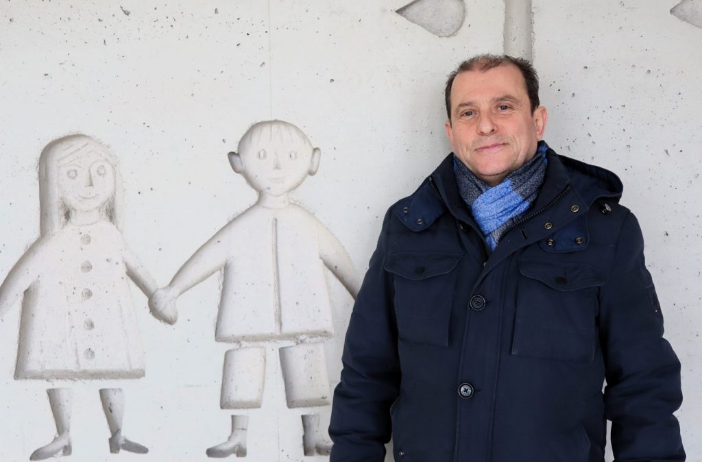 Hausmeister, Mann vor Wand mit Kinderzeichnungen und Winterbekleidung