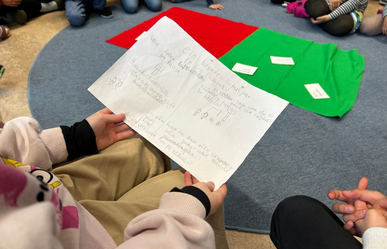 Kinder sitzen in einem Sitzkreis um einem blauen Teppich. Dort liegen zwei Tücher in rot und grün. Ein Kind ist von hinten zu sehen wie es ein Blatt mit Vorschlägen für die Klassenkonferenz in der Hand hält.